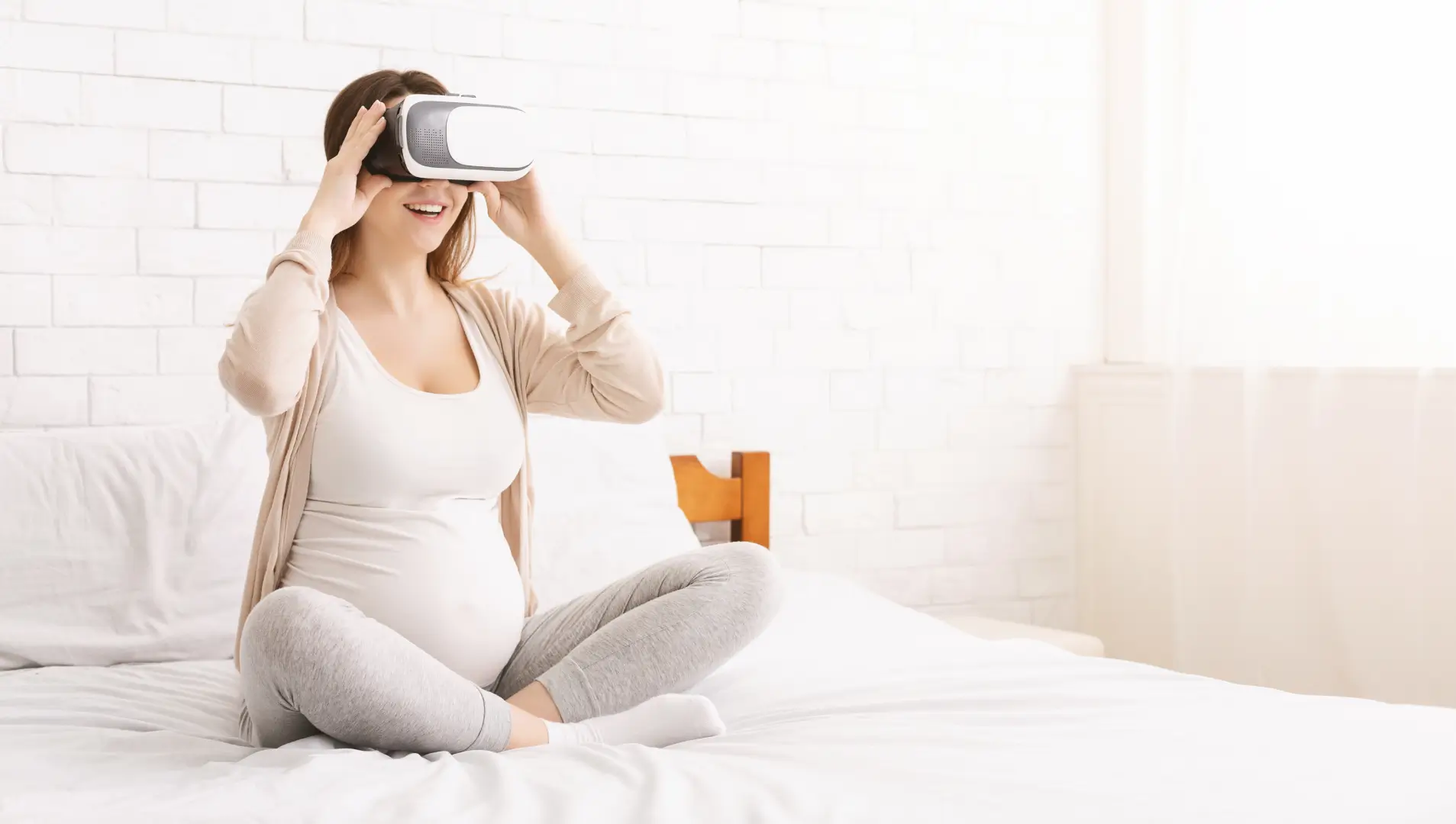 Eine schwangere Frau sitzt auf einem Bett. Sie trägt eine VR-Brille und lacht.