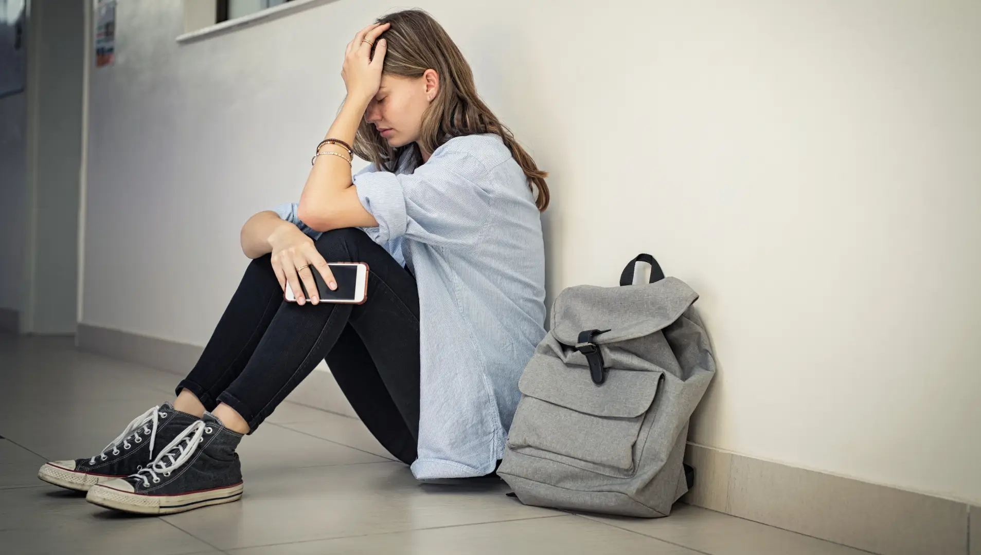 Auf dem Bild sitzt eine junge Frau mit einem traurigen Gesichtsausdruck auf dem Boden eines Flurs, während sie ein Smartphone in der Hand hält. Neben ihr steht ein grauer Rucksack.