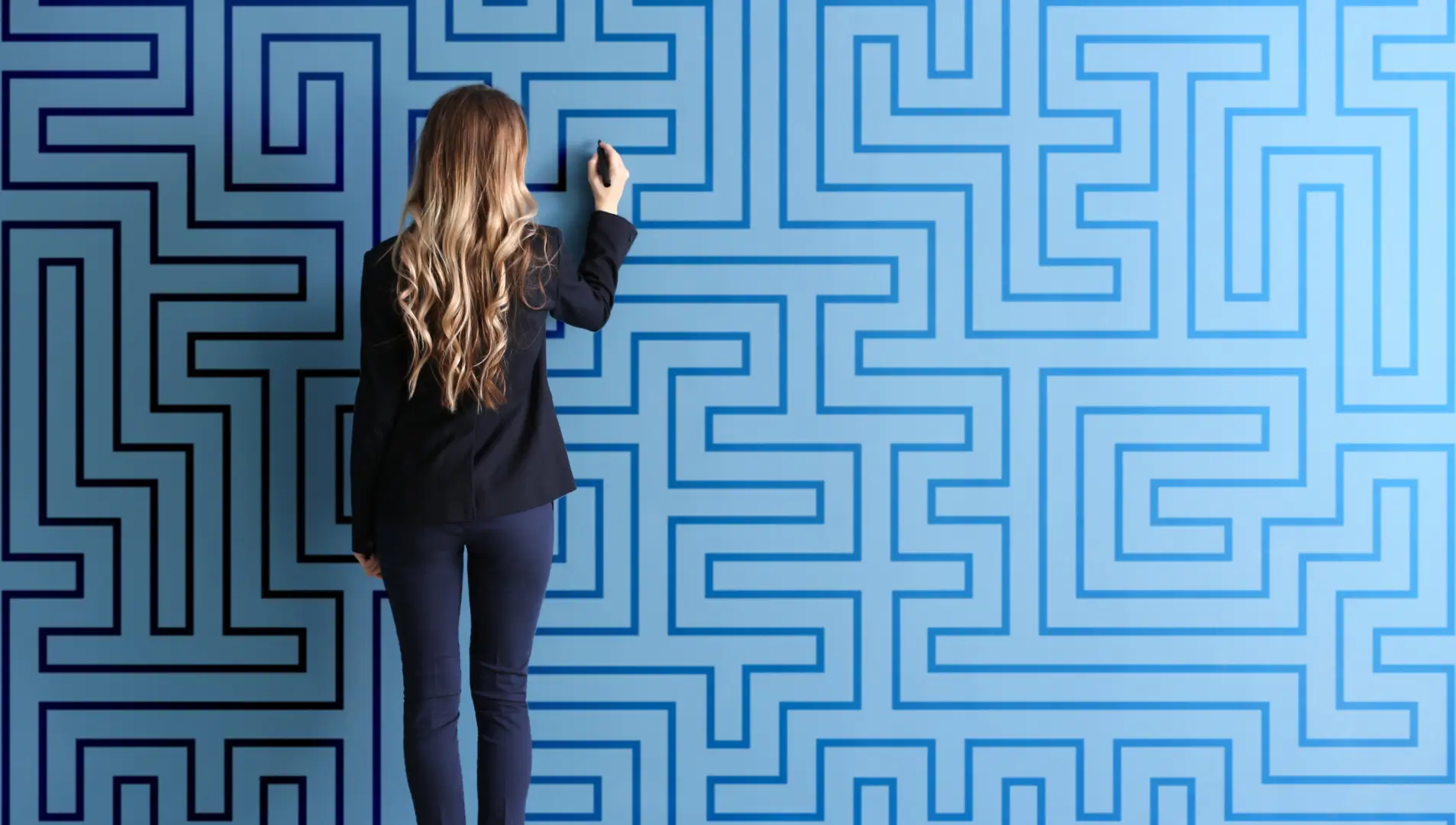 Auf dem Bild steht eine Frau mit langen, welligen Haaren in Geschäftskleidung vor einer großen blauen Wand, die mit einem Labyrinthmuster bedeckt ist. Sie scheint mit einem Stift auf die Wand zu zeichnen oder eine Lösung für das Labyrinth zu suchen.