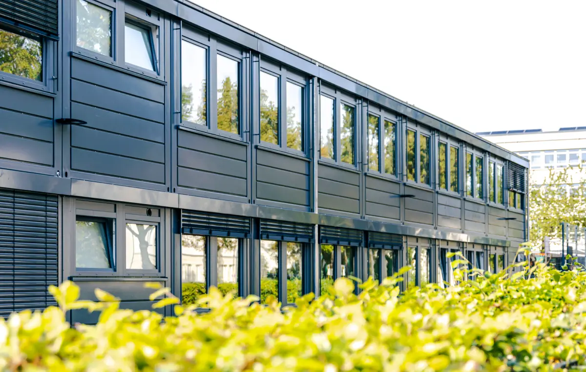 Ein modernes, zweistöckiges Gebäude mit großen Fenstern und grauen Paneelen ist zu sehen. Im Vordergrund befindet sich eine grüne Hecke, die das Gebäude teilweise verdeckt. Der Himmel ist klar und sonnig.