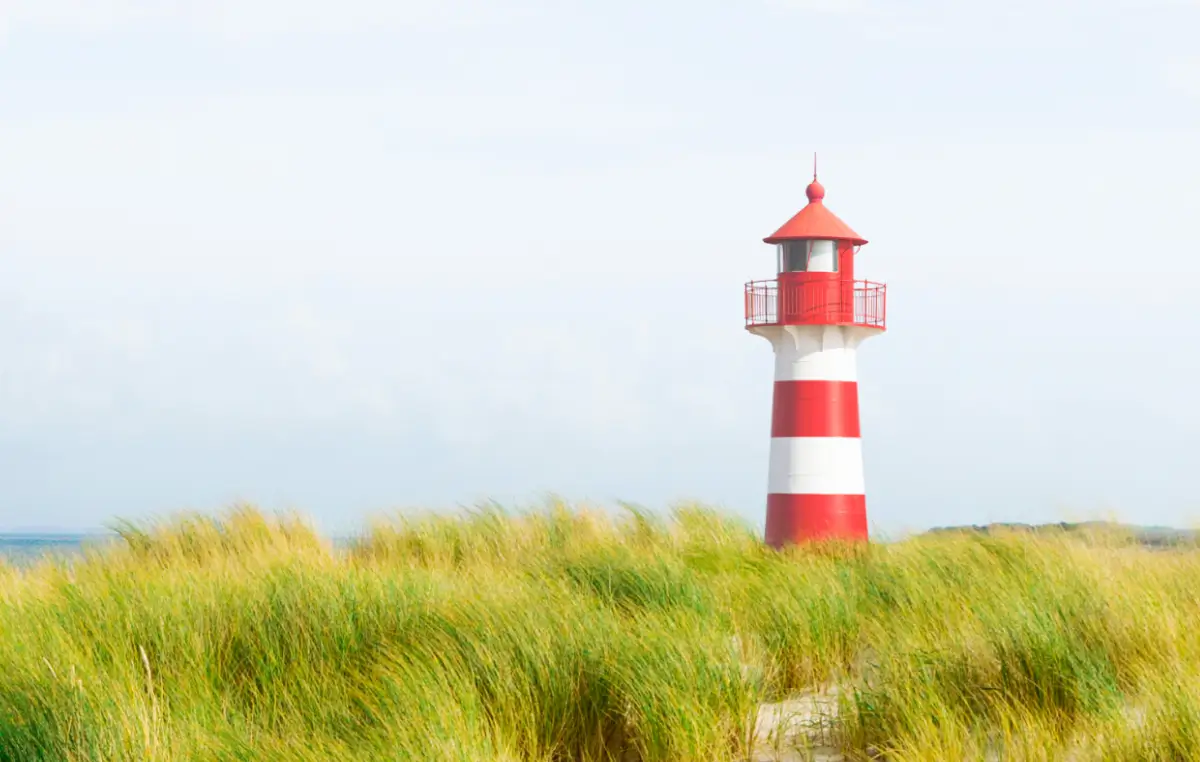 Ein rot-weiß gestreifter Leuchtturm steht hinter Dünen am Meer unter einem hellen Himmel.