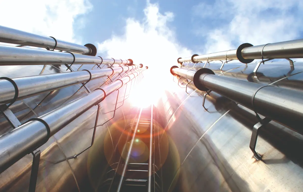 Blick nach oben auf mehrere große, glänzende Rohre mit Sonnenlicht im Hintergrund.