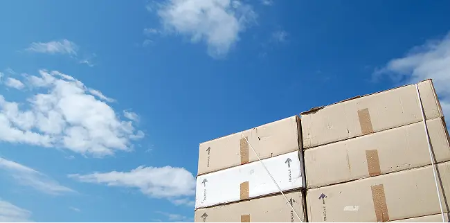 Mehrere große Kartons sind zu einem Stapel übereinandergeschichtet und mit Band gesichert. Darüber ist ein blauer Himmel mit vereinzelten Wolken zu sehen.