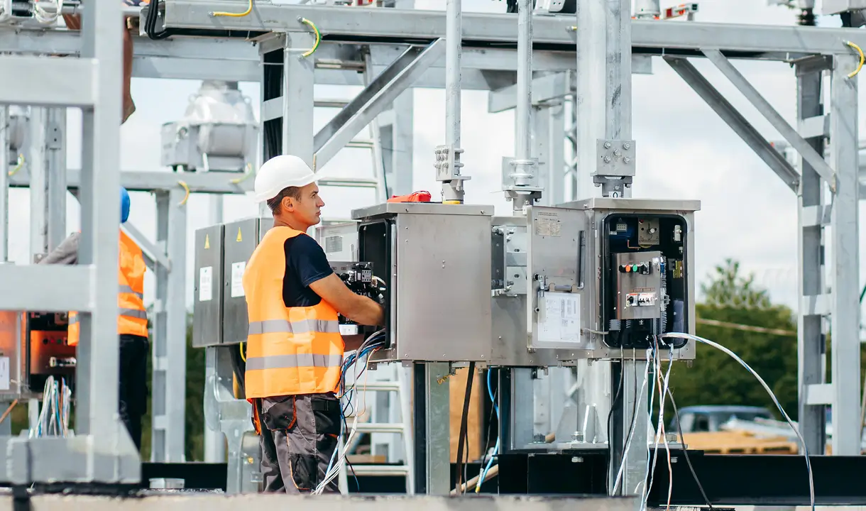 Ein Arbeiter mit Helm und Warnweste arbeitet an einem offenen Schaltschrank auf einem Umspannwerksgelände mit Metallkonstruktionen und technischen Anlagen im Freien.