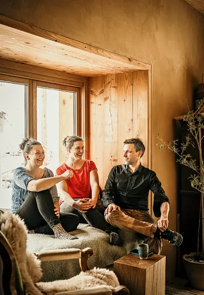 Alexandra Schenker-Primus, Sarah Hoppe und Florian Hoppe sitzen lachend und entspannt auf einer gemütlichen Sitzbank in einem warmen, holzverkleideten Raum vor einem Fenster.