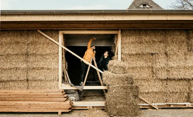 Das Bild zeigt zwei Personen, die an einem Gebäude aus Strohballen arbeiten, wobei eine auf einer Leiter steht und die andere daneben steht.