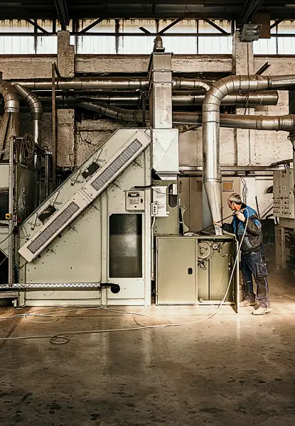 Eine Person in Arbeitskleidung bedient eine große industrielle Maschine in einer Fabrikumgebung mit zahlreichen Rohren und Förderbändern.