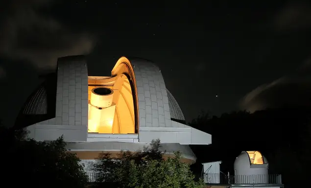 Das Bild zeigt zwei beleuchtete Kuppeln eines Observatoriums bei Nacht, wobei die größere der beiden Kuppeln ein großes Teleskop enthüllt.