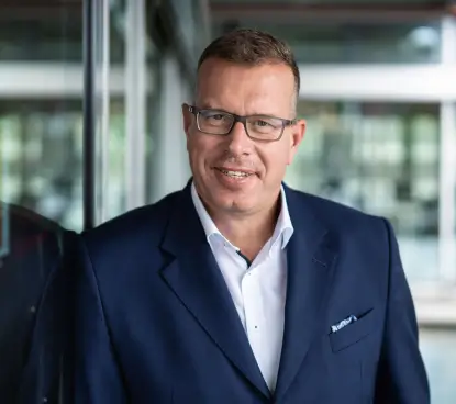 Prof. Andreas Tünnermann in einem dunkelblauen Anzug und Brille lächelt in die Kamera und lehnt an einer Glaswand in einem modernen Bürogebäude.