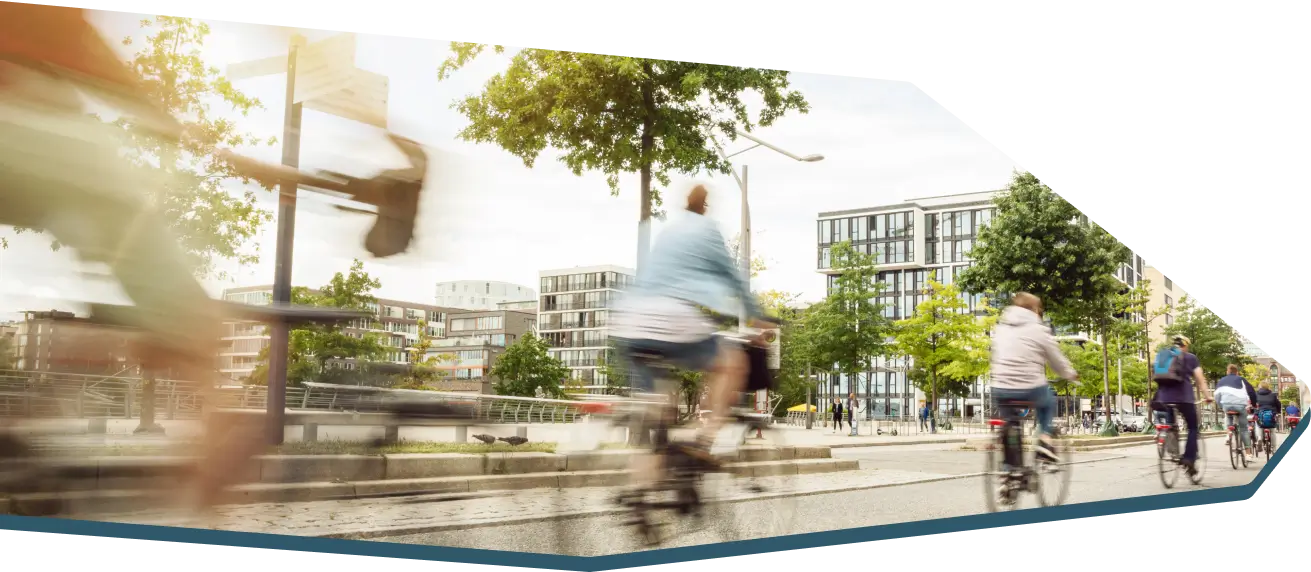 Das Bild zeigt eine städtische Szene mit mehreren Menschen, die mit Fahrrädern auf einer Straße fahren, vor modernen Gebäuden und Bäumen.