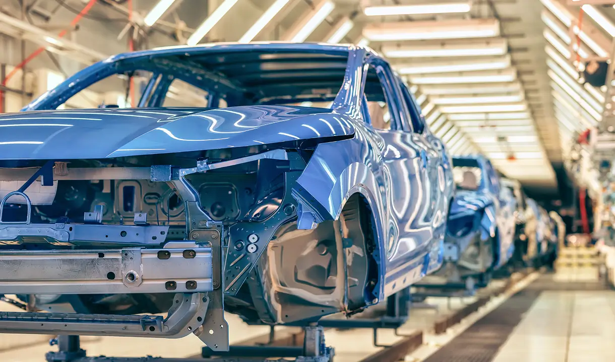 Eine blaue Auto-Karosserie bewegt sich durch eine Produktionslinie in einer hell beleuchteten Fabrikhalle.