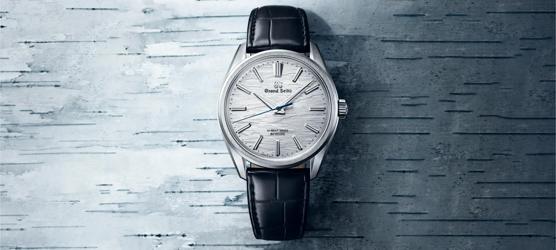 Diese Grand Seiko Uhr hebt sich durch ihr silbernes, texturiertes Zifferblatt, einen blauen Sekundenzeiger und ein schwarzes Lederarmband vor einem passenden Hintergrund hervor.