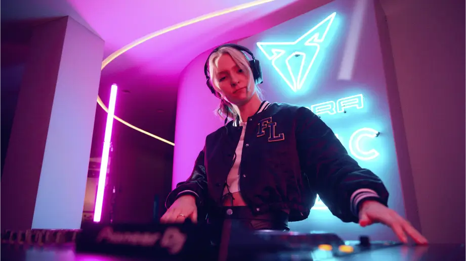Eine Person mit Kopfhörern steht an einem DJ-Pult, in eine Jacke gekleidet, und wirkt konzentriert auf die Musik. Im Hintergrund leuchtet ein farbenfrohes Neonlicht, das eine moderne und energiegeladene Atmosphäre erzeugt.