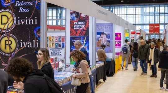 Besucher schlendern durch die Gänge einer Buchmesse, umgeben von Ständen mit bunten Buchplakaten und ausgestellten Büchern. Die Atmosphäre ist lebhaft und interessiert.