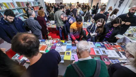 Menschen stöbern auf einer Buchmesse an einem mit Büchern gefüllten Stand. Der Raum ist gut besucht, und die Besucher schauen sich interessiert die ausgestellten Bücher an.