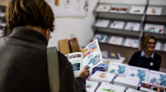 Personen blättern durch Bücher auf einem Tisch. Eine Person im karierten Mantel hält ein aufgeschlagenes Buch, während andere ebenfalls Bücher betrachten. Die Szene wirkt aufmerksam und interessiert.