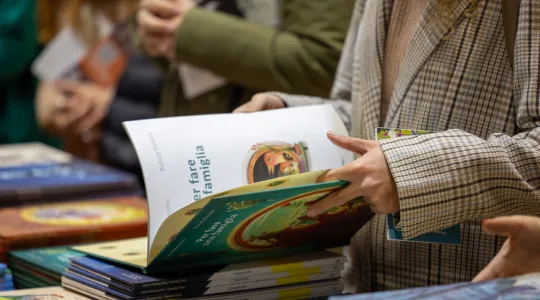 Menschen blättern in Büchern bei einer Buchmesse. Eine Person hält mehrere bunte Bücher in den Händen, während andere im Hintergrund ebenfalls Bücher betrachten. Die Szene vermittelt eine neugierige und interessierte Atmosphäre.