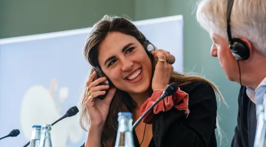 Eine Frau lächelt fröhlich und trägt Kopfhörer, während sie an einem Tisch mit Mikrofonen sitzt. Neben ihr sitzt ein Mann, der ebenfalls Kopfhörer trägt. Die Szene wirkt entspannt und kommunikativ.