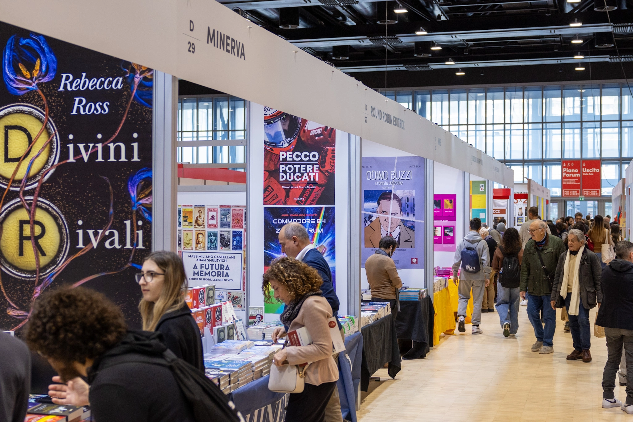 Besucher schlendern durch die Gänge einer Buchmesse, umgeben von Ständen mit bunten Buchplakaten und ausgestellten Büchern. Die Atmosphäre ist lebhaft und interessiert.