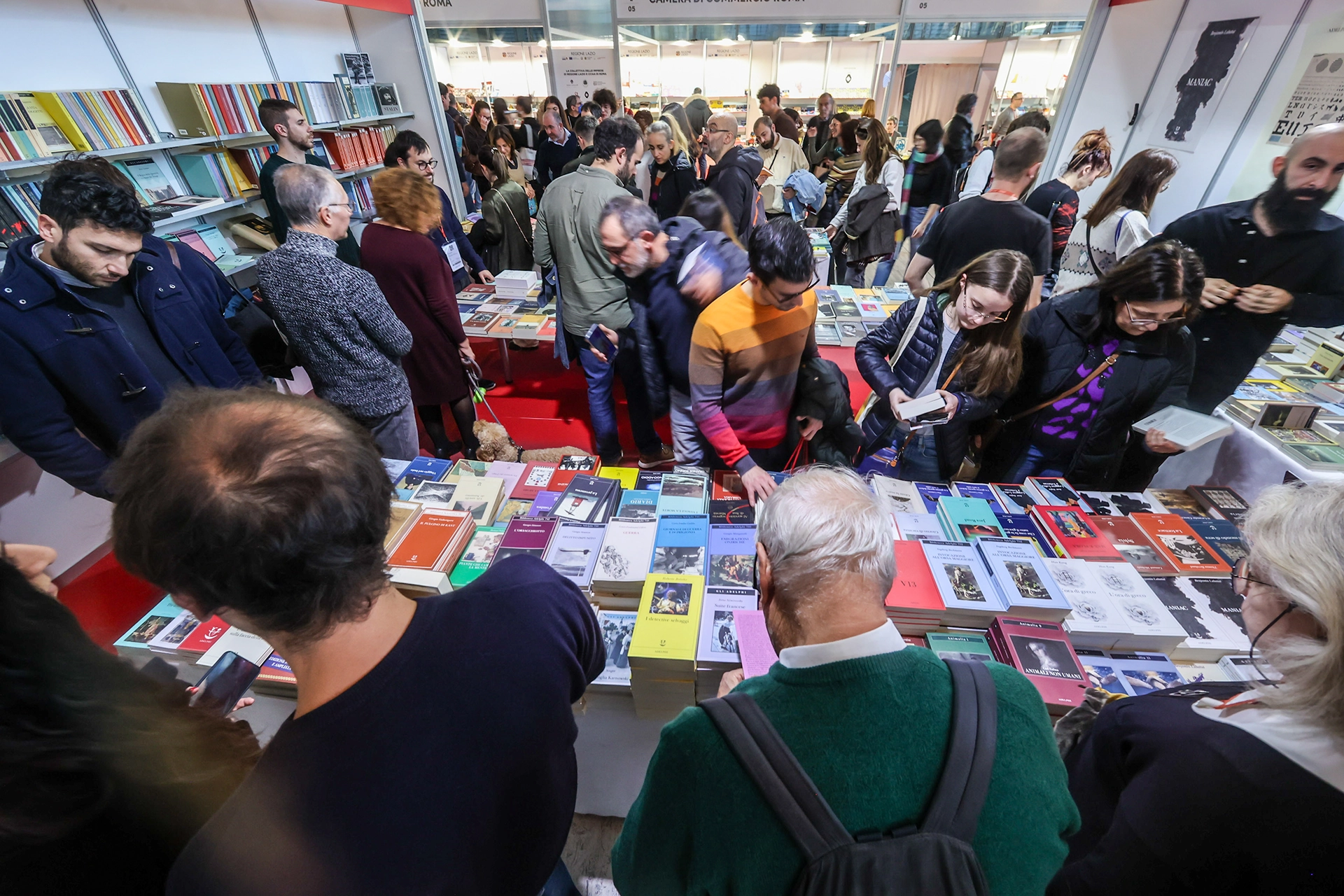 Menschen stöbern auf einer Buchmesse an einem mit Büchern gefüllten Stand. Der Raum ist gut besucht, und die Besucher schauen sich interessiert die ausgestellten Bücher an.