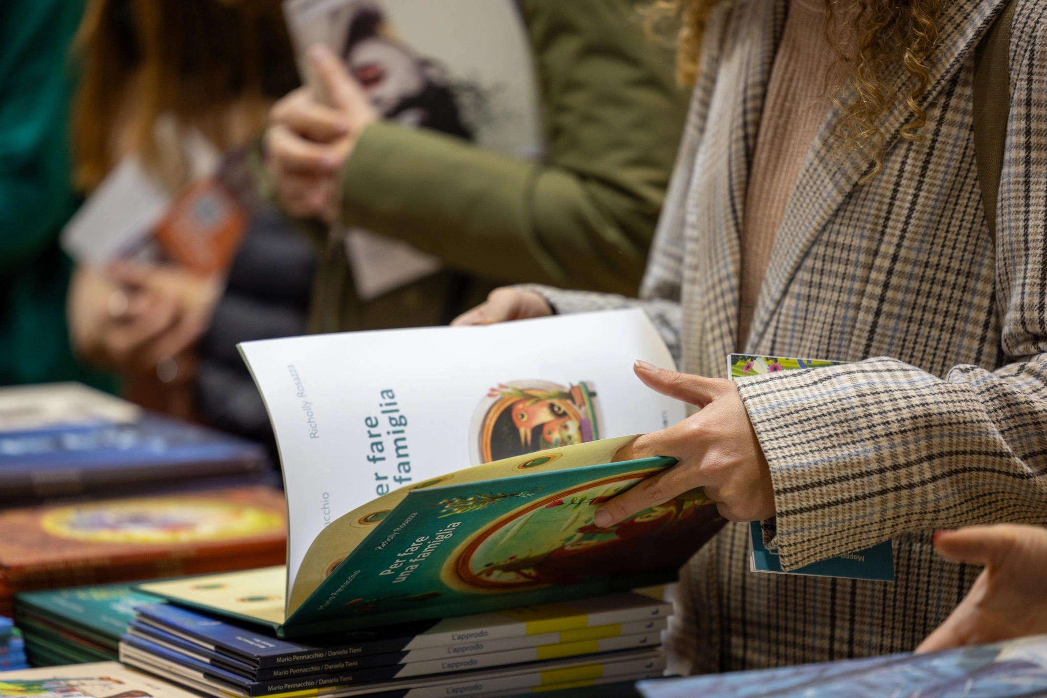 Menschen blättern in Büchern bei einer Buchmesse. Eine Person hält mehrere bunte Bücher in den Händen, während andere im Hintergrund ebenfalls Bücher betrachten. Die Szene vermittelt eine neugierige und interessierte Atmosphäre.