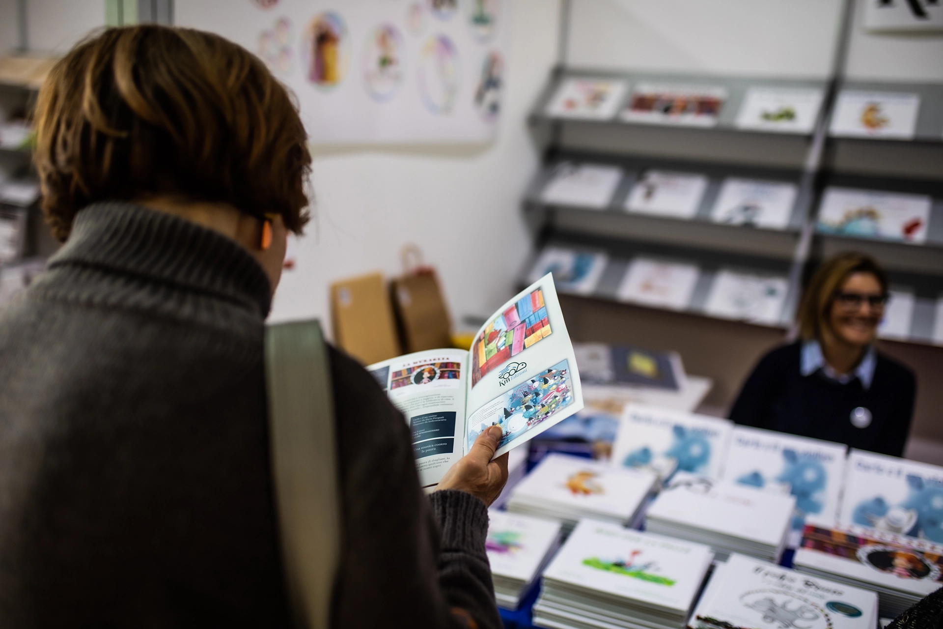 Personen blättern durch Bücher auf einem Tisch. Eine Person im karierten Mantel hält ein aufgeschlagenes Buch, während andere ebenfalls Bücher betrachten. Die Szene wirkt aufmerksam und interessiert.
