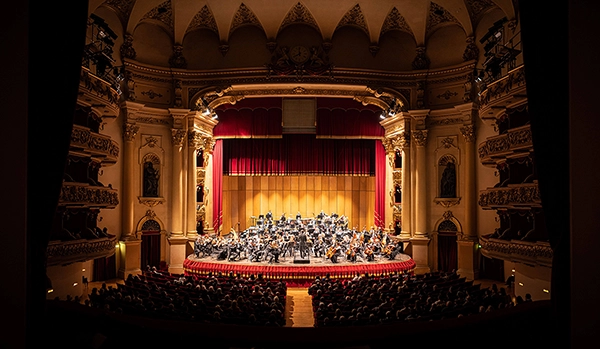 Ein Orchester spielt auf der Bühne eines prächtigen, reich verzierten Konzertsaals. Der Saal ist mit roten Vorhängen und goldenen Dekorationen geschmückt. Viele Menschen sitzen im Publikum und verfolgen das Konzert.