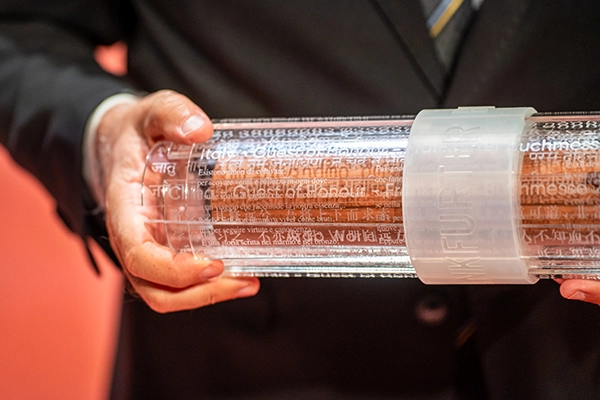 Eine Person hält eine transparente Rolle mit eingravierten Texten in verschiedenen Sprachen. Die Rolle ist mit einem Band gesichert, auf dem 'Frankfurter Buchmesse' steht. Der Hintergrund ist unscharf und in warmen Tönen gehalten.