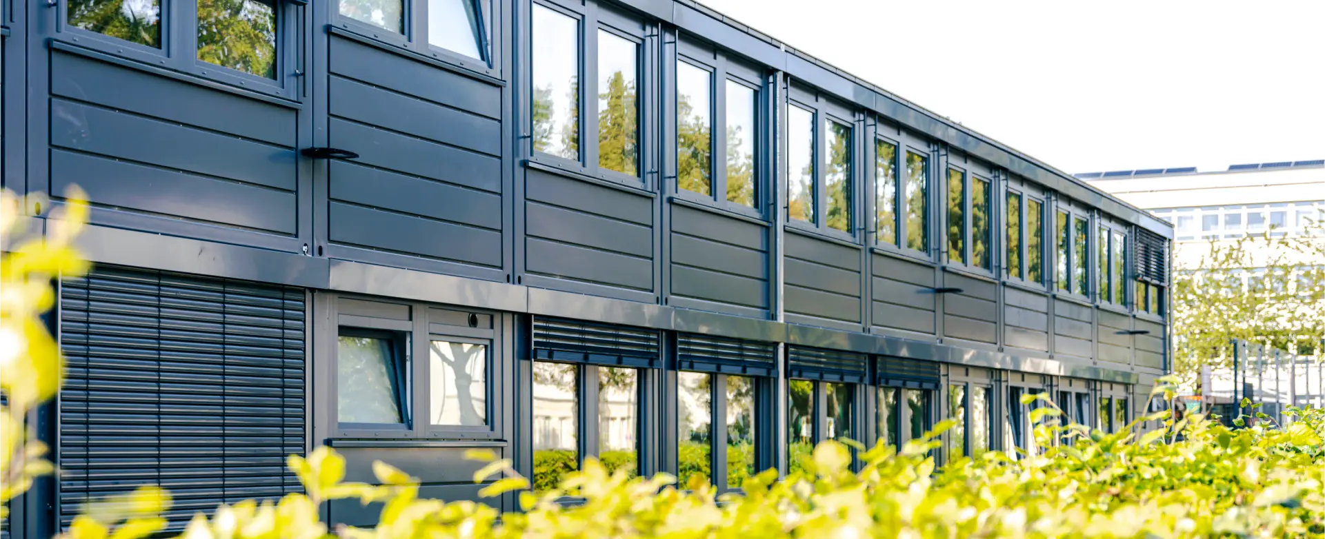 Ein modernes, zweistöckiges Gebäude mit großen Fenstern und grauen Paneelen ist zu sehen. Im Vordergrund befindet sich eine grüne Hecke, die das Gebäude teilweise verdeckt. Der Himmel ist klar und sonnig.