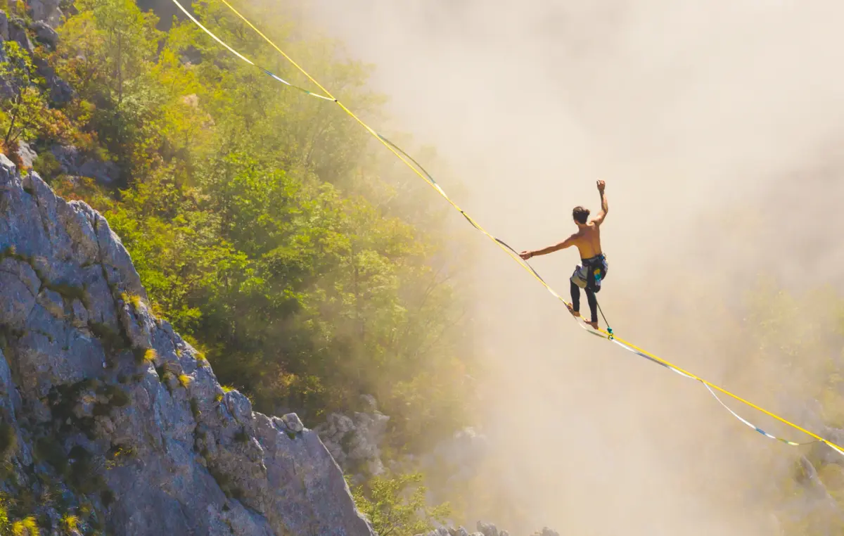 Eine Person balanciert auf einer Slackline, die über eine Schlucht zwischen Felsen gespannt ist. Die Umgebung ist von grünen Bäumen und Felsen geprägt, und Nebel umhüllt die Szene teilweise. Die Person ist oberkörperfrei und trägt Kletterausrüstung.