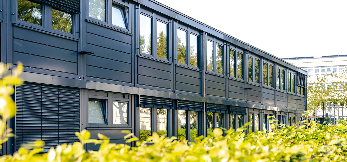 Ein modernes, zweistöckiges Gebäude mit großen Fenstern und grauen Paneelen ist zu sehen. Im Vordergrund befindet sich eine grüne Hecke, die das Gebäude teilweise verdeckt. Der Himmel ist klar und sonnig.