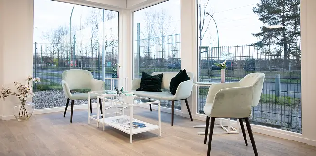 Ein heller Warteraum mit großen Fenstern, die den Blick nach draußen auf einen Zaun und Bäume freigeben. Der Raum ist mit hellen Polstersesseln, einem kleinen Sofa mit dunklen Kissen, einem weißen Couchtisch und dekorativen Blumen ausgestattet. Der Boden ist aus hellem Holz.