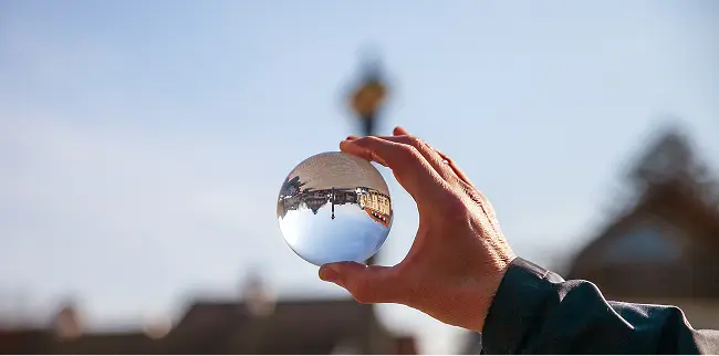 Eine Hand hält eine glasklare Kugel, die eine umgedrehte Spiegelung einer städtischen Szenerie zeigt. Im Hintergrund ist unscharf ein Gebäude zu sehen, während der Himmel blau ist.