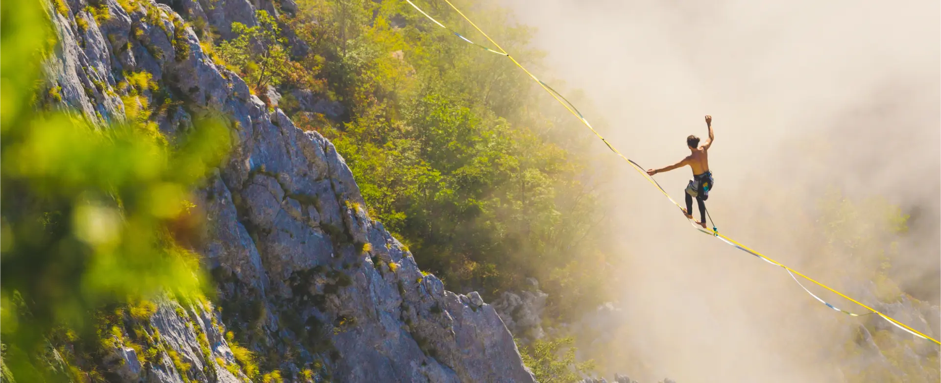 Eine Person balanciert auf einer Slackline, die über eine Schlucht zwischen Felsen gespannt ist. Die Umgebung ist von grünen Bäumen und Felsen geprägt, und Nebel umhüllt die Szene teilweise. Die Person ist oberkörperfrei und trägt Kletterausrüstung.