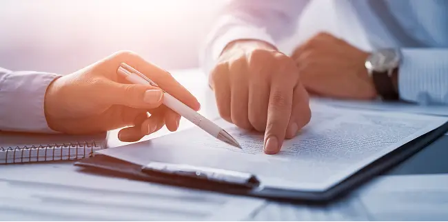 Zwei Personen betrachten ein Dokument. Eine Hand hält einen Stift, während die andere auf den Text zeigt. Beide tragen Hemden, und die Szene ist in warmes Licht getaucht.