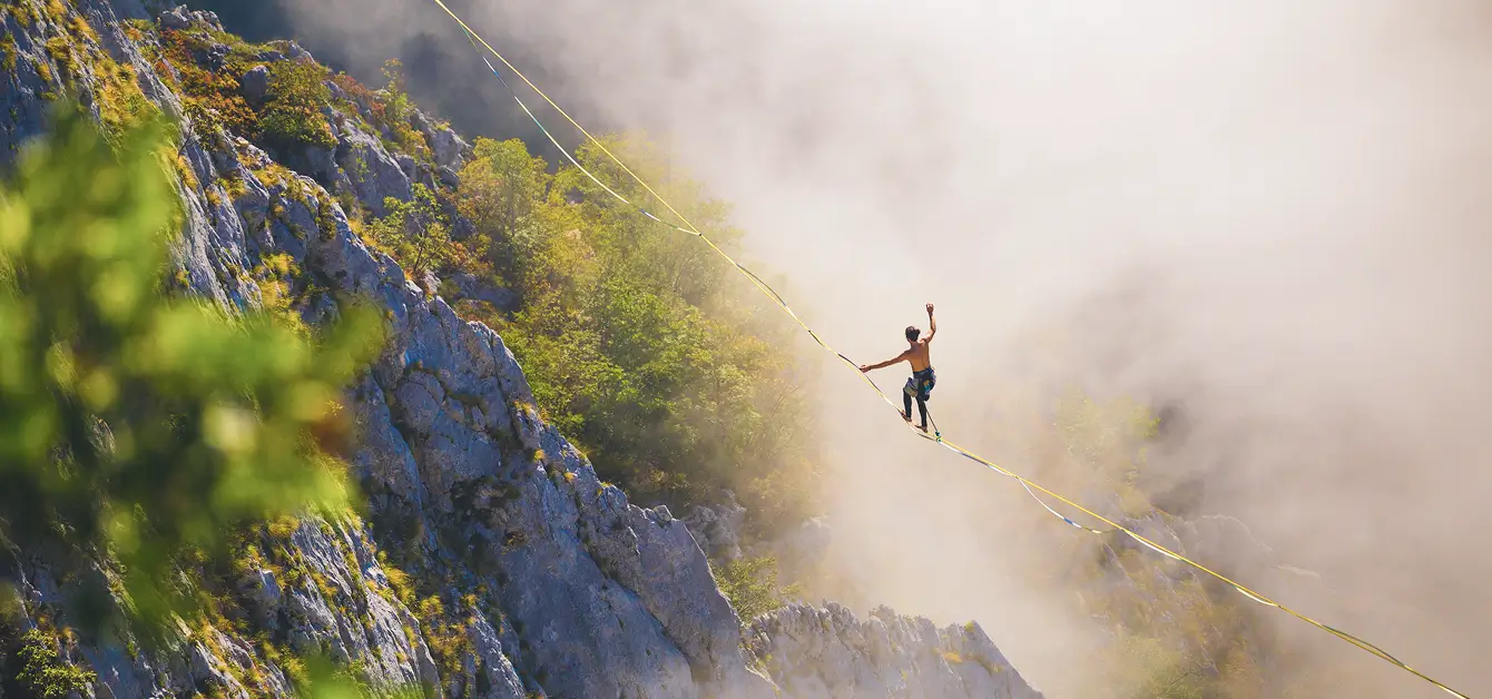 Eine Person balanciert auf einer Slackline, die über eine Schlucht zwischen Felsen gespannt ist. Die Umgebung ist von grünen Bäumen und Felsen geprägt, und Nebel umhüllt die Szene teilweise. Die Person ist oberkörperfrei und trägt Kletterausrüstung.