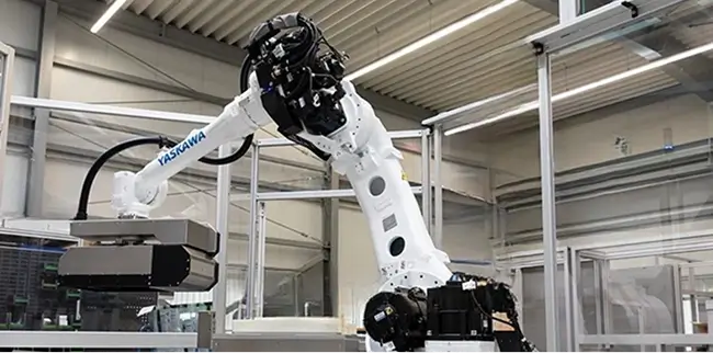 Ein industrieller Roboterarm der Marke Yaskawa in einer Fabrikumgebung. Der Arm ist weiß und mit verschiedenen mechanischen Komponenten ausgestattet. Im Hintergrund sind Metallrahmen und Fabrikbeleuchtung zu sehen.