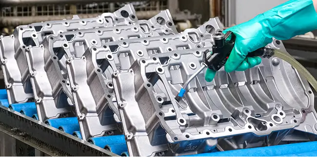 Eine Hand mit grünen Handschuhen reinigt oder bearbeitet präzise eine Reihe von metallischen Gussteilen mit einem Druckluftwerkzeug.