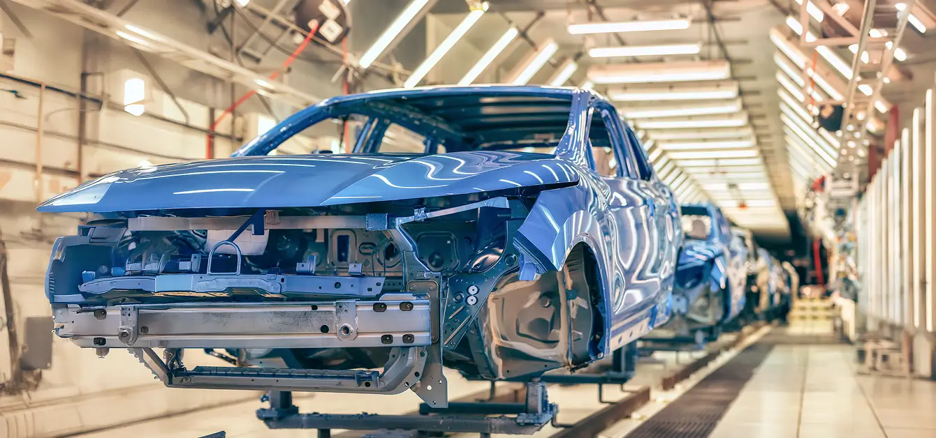 Eine blaue Auto-Karosserie bewegt sich durch eine Produktionslinie in einer hell beleuchteten Fabrikhalle.