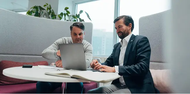 Zwei Personen sitzen an einem Tisch mit einem Laptop, Papieren und einem offenen Notizbuch und besprechen gemeinsam ein Projekt.
