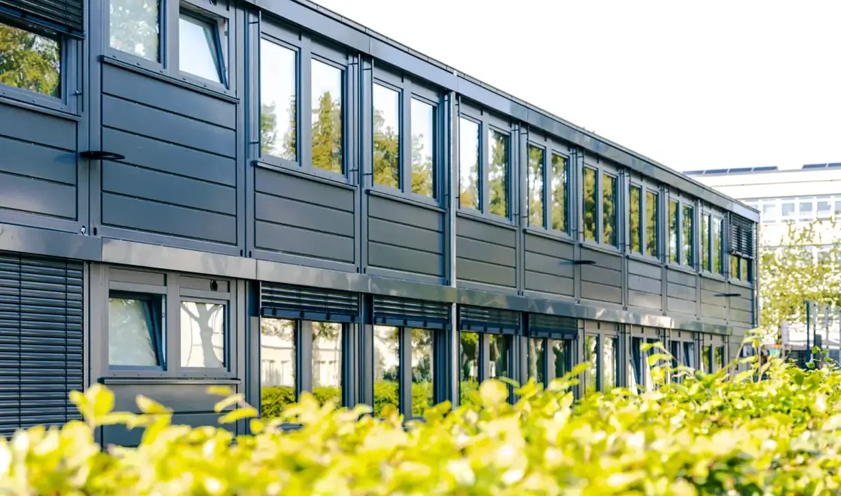 Ein modernes, zweistöckiges Gebäude mit großen Fenstern und grauen Paneelen ist zu sehen. Im Vordergrund befindet sich eine grüne Hecke, die das Gebäude teilweise verdeckt. Der Himmel ist klar und sonnig.