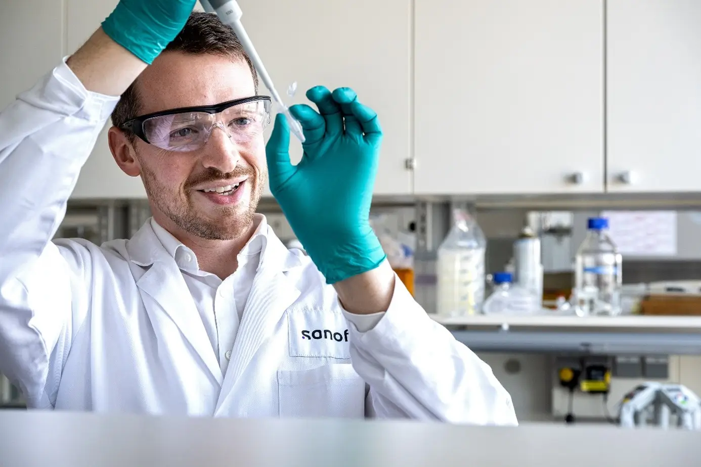 Wissenschaftler im Labor hält eine Pipette und eine kleine Probe, trägt grüne Schutzhandschuhe und arbeitet in einer professionellen Laborumgebung mit verschiedenen Chemikalien und Geräten im Hintergrund.