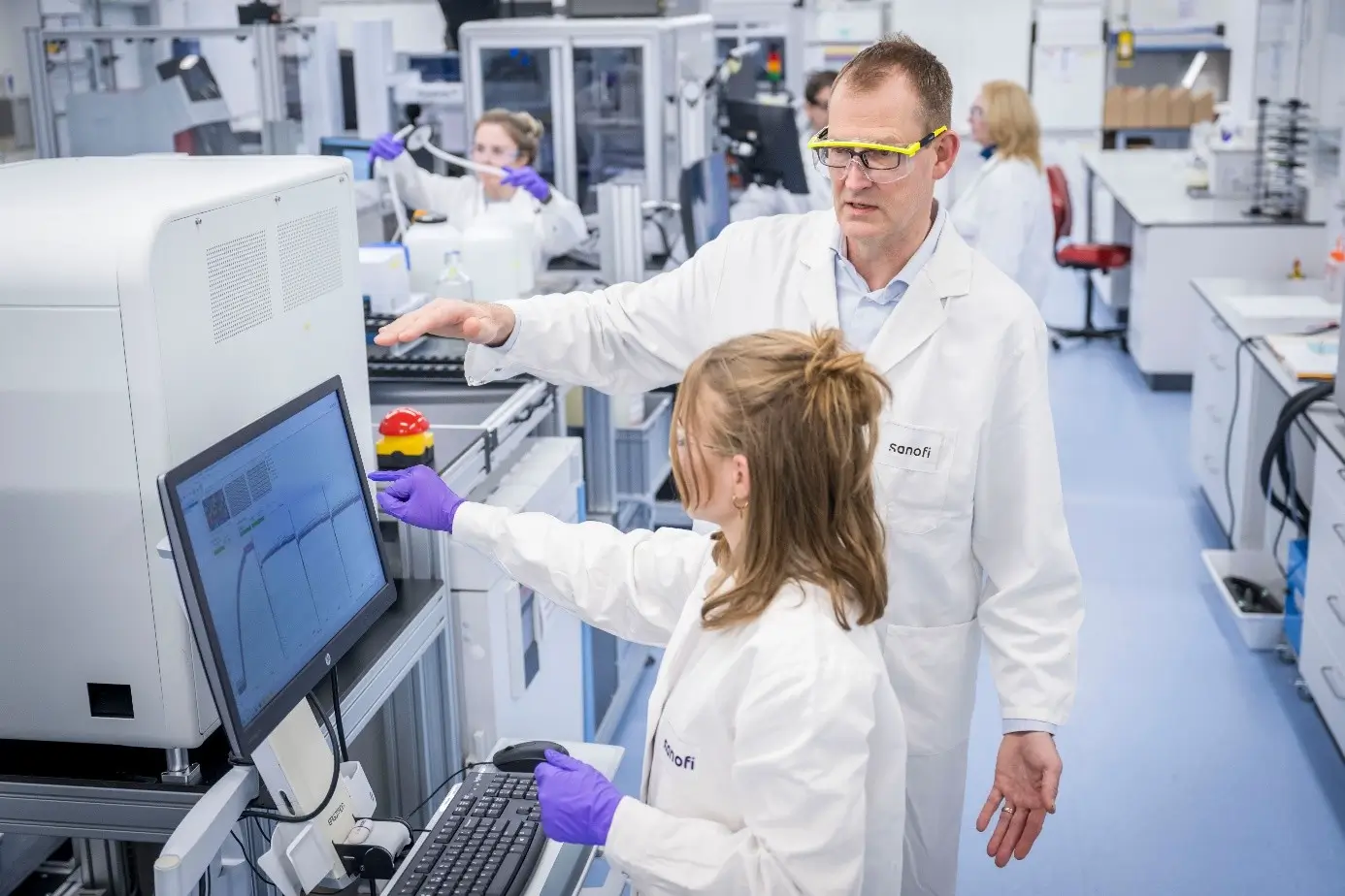 Zwei Wissenschaftler im Labor analysieren Daten auf einem Computerbildschirm, während sie von fortschrittlicher Laborausrüstung und Arbeitsstationen umgeben sind.