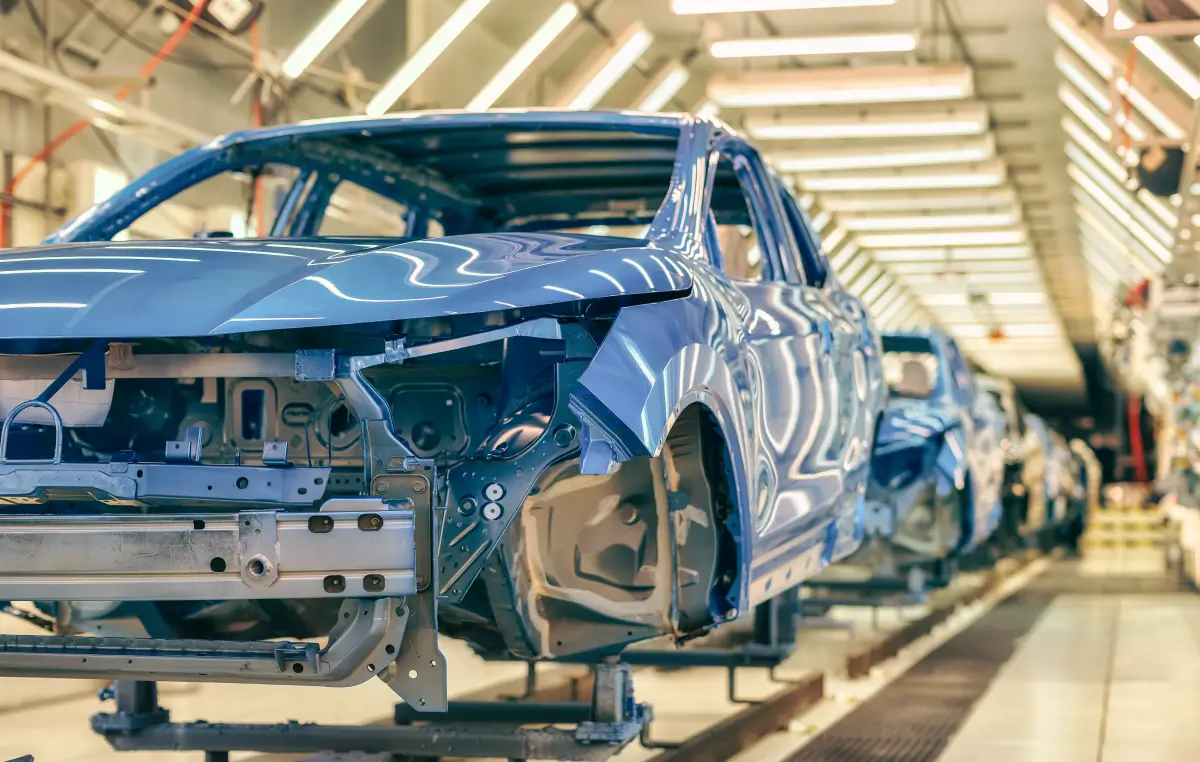 Eine blaue Auto-Karosserie bewegt sich durch eine Produktionslinie in einer hell beleuchteten Fabrikhalle.