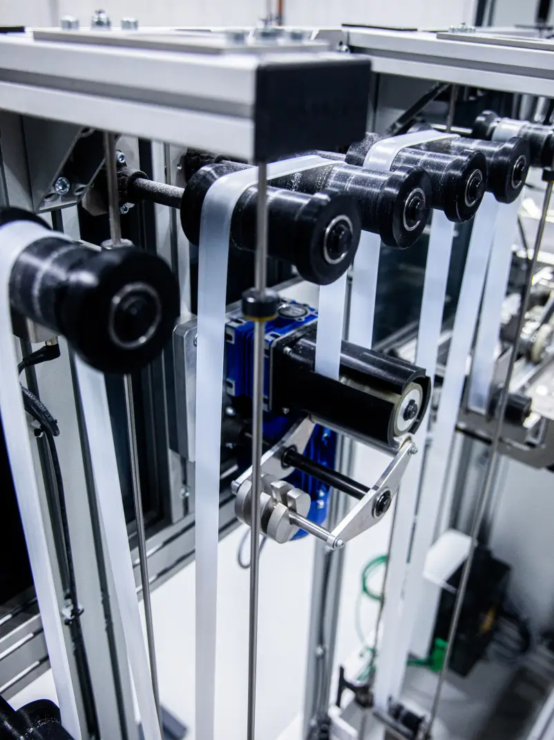 Eine Maschine mit mehreren schwarzen Rollen, durch die transparente Fahrradschläuche geführt werden. Die Schläuche verlaufen parallel durch die Mechanik, die aus Metall- und Kunststoffteilen besteht. Im Hintergrund sind weitere technische Komponenten sichtbar.
