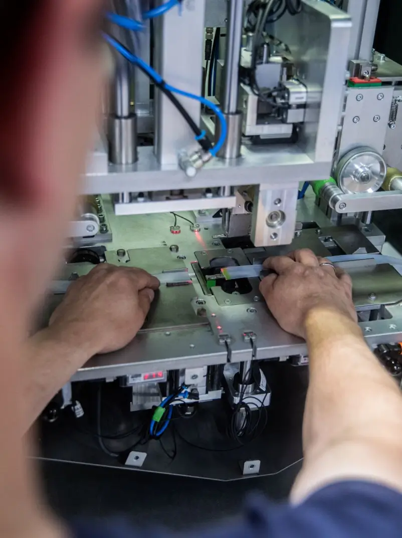 Eine Person arbeitet an einer präzisen Maschine mit metallischen und elektronischen Komponenten. Die Hände platzieren ein Materialstück in die Maschine, während blaue Kabel und technische Details im Aufbau erkennbar sind.
