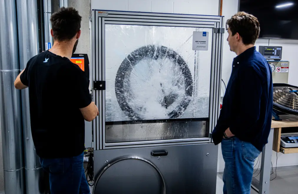 Zwei Personen stehen vor einer Maschine, die einen Fahrradreifen in einem Testprozess zeigt. Wasser spritzt gegen eine durchsichtige Schutzscheibe, während die Männer die Szene beobachten.