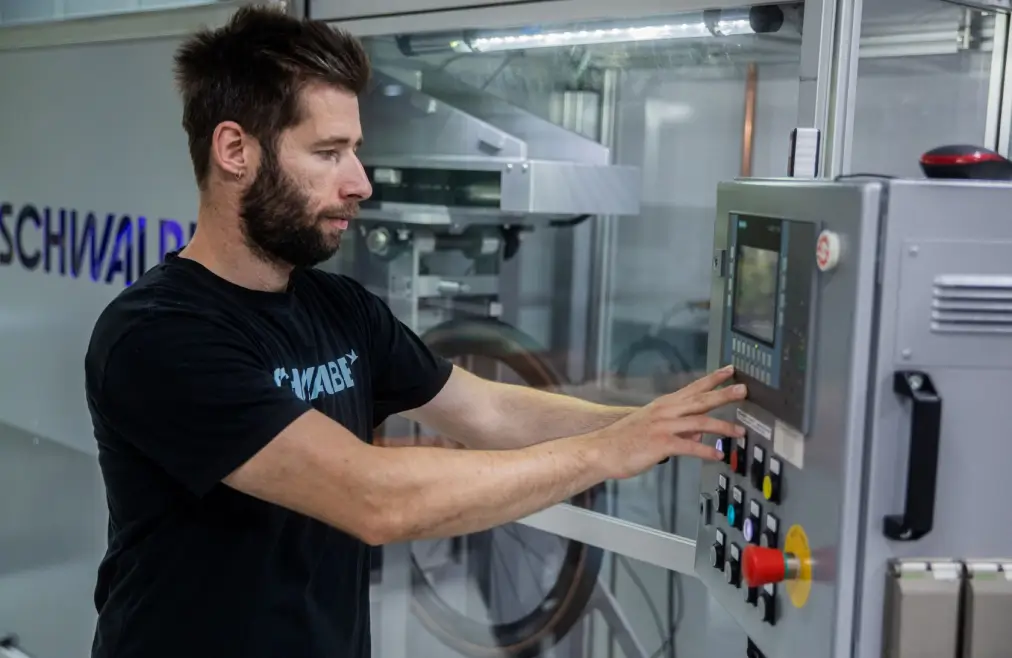 Eine Person bedient ein Bedienfeld mit mehreren Knöpfen und einem Display. Im Hintergrund steht eine Maschine mit der Aufschrift „Schwalbe“, und ein Fahrradreifen ist teilweise sichtbar.