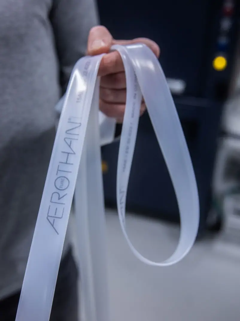 Eine Hand hält mehrere transparente Fahrradschläuche mit der Aufschrift „Aerothan“. Die Schläuche sind flexibel und leicht glänzend. Der Hintergrund ist unscharf und zeigt eine industrielle Umgebung.