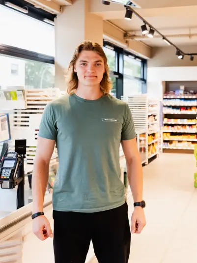 Ein junger Mann steht in einem modernen Supermarkt an der Kasse. Er trägt ein olivgrünes T-Shirt und eine schwarze Hose. Im Hintergrund sind Regale mit Produkten und große Fenster zu sehen.